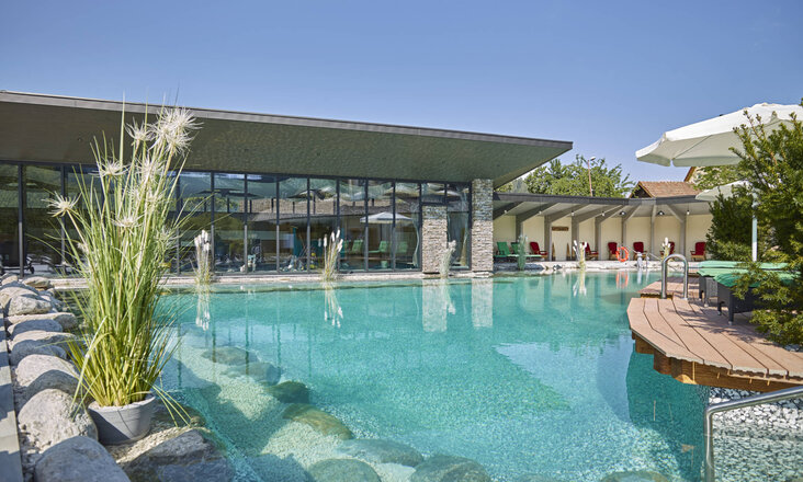Large swimming pond with Balena system and plants at the “Heselbacher Hof” wellness hotel in Baiersbronn, with the hotel in the background