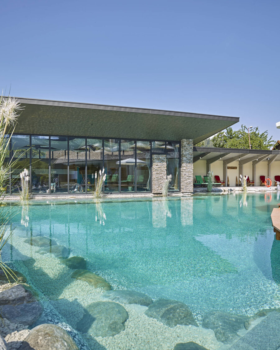 Large swimming pond with Balena system and plants at the “Heselbacher Hof” wellness hotel in Baiersbronn, with the hotel in the background