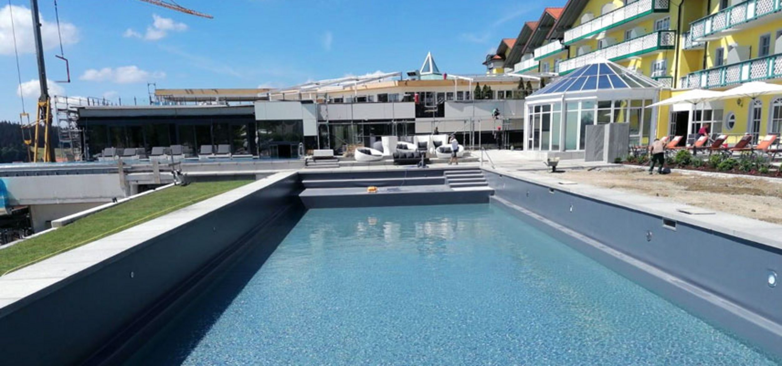 View from the natural pool over the pool stairs to the main building of the natural pool with loungers and guests at the sports and wellness hotel “Angerhof” in St. Englmar
