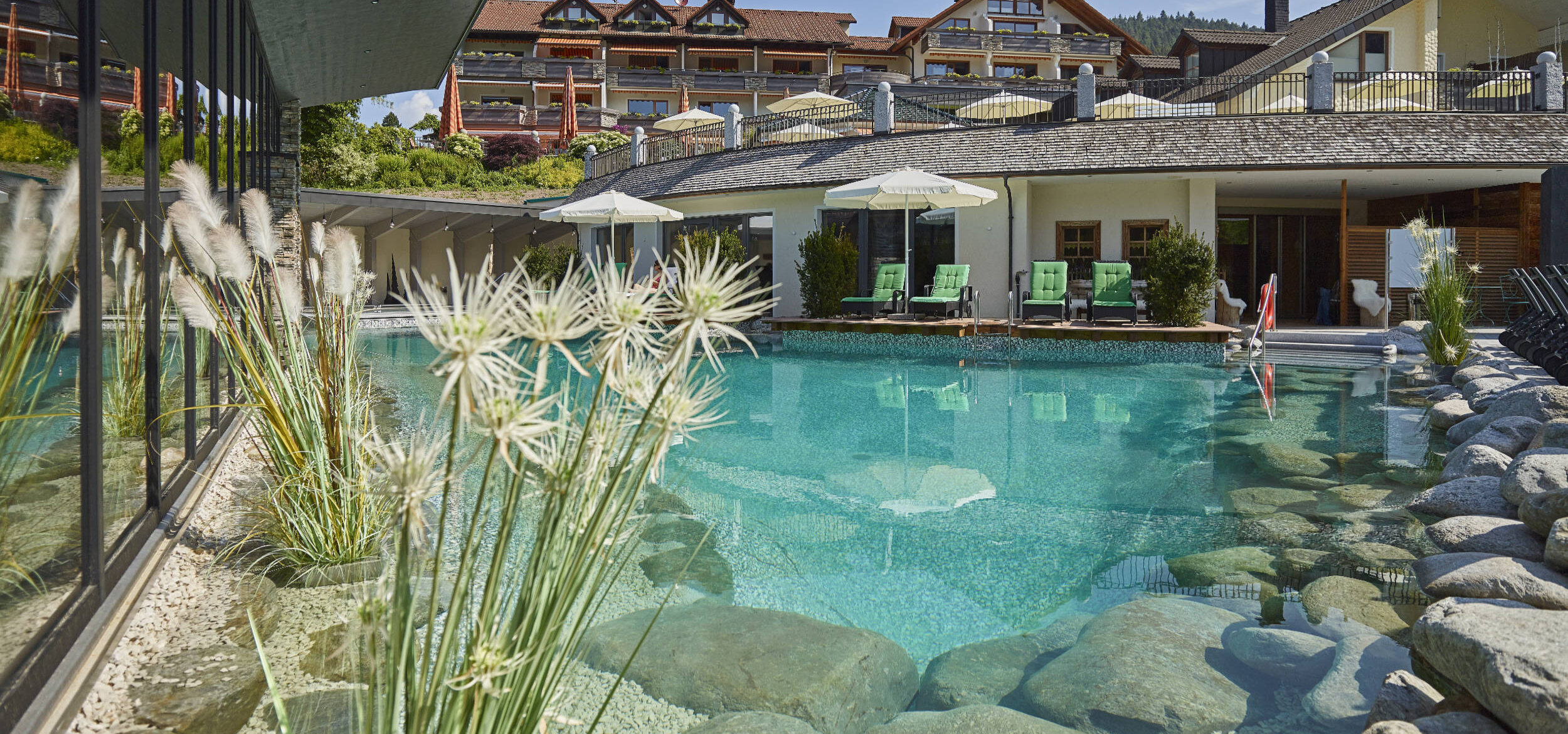 Natural swimming pond with Balena system at the “Heselbacher Hof” wellness hotel in Baiersbronn with greenery and stones at the edge, deckchairs in the background